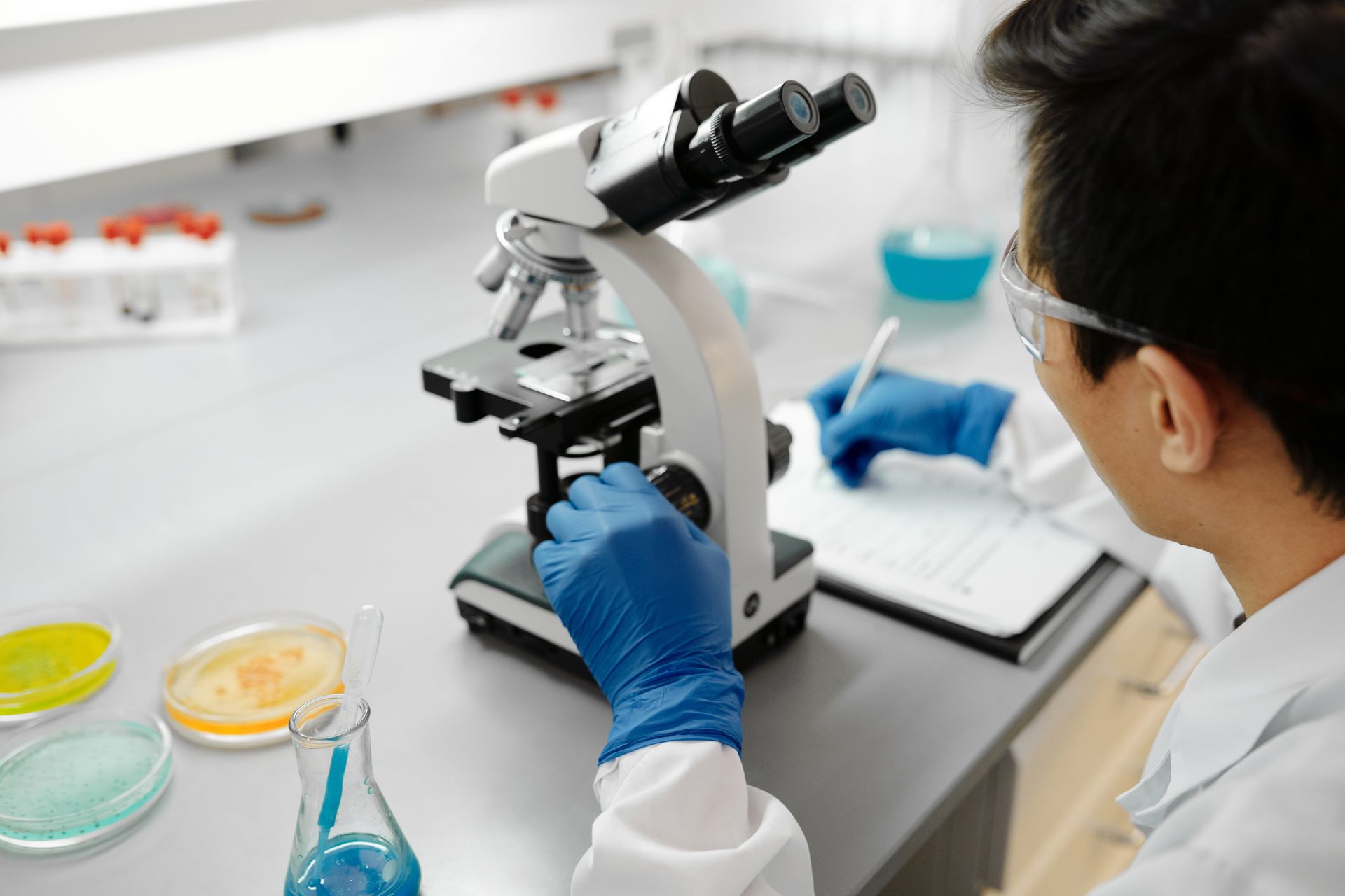 A laboratory technician examining a specimen under a microscope beside sample dishes